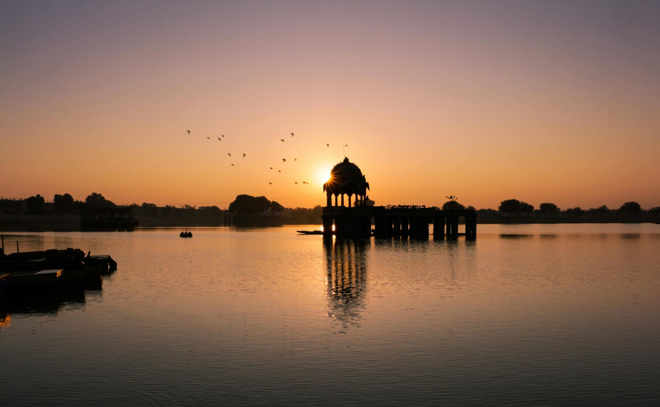 A scenic sunset over a calm lake, featuring a silhouetted temple and flying birds.
